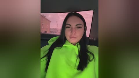 Video of a young woman with long black hair, wearing a bright green hoodie, standing in a dimly lit room with a large TV displaying a blurry image of a brick building in the background.