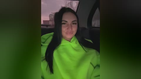 A video of a young woman with long, black hair, wearing a bright green hoodie, seated in a dimly lit car. The background shows a blurred cityscape.