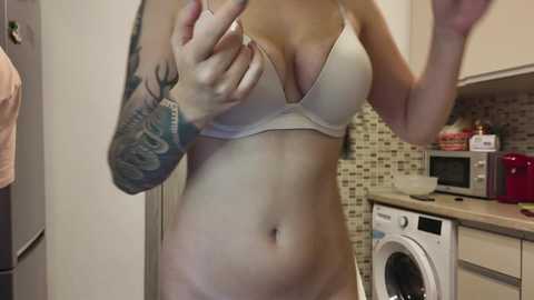 Video of a woman with light skin, tattoos, and a white bra, standing in a modern kitchen with beige tiles, white appliances, and a red toaster.