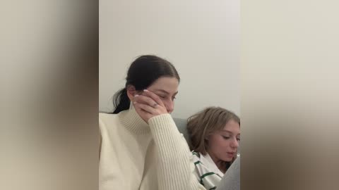 Media: Video of two girls sitting in a classroom, one with a white sweater, the other with a green and white striped shirt, both looking down, in a dimly lit room with soft focus.