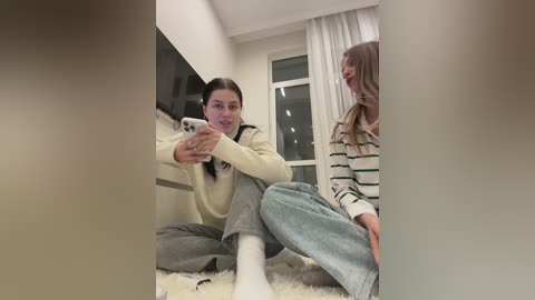 Media: Video of two young women, one in a beige sweater and grey pants, the other in a striped sweater, sitting on a white carpet in a cozy room with a window and white curtains.