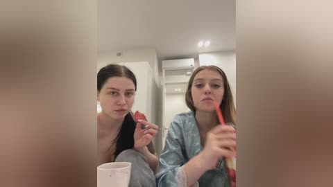 Media: A video of two young women, one with dark hair, one with light hair, both in robes, brushing teeth, in a modern, white-tiled bathroom.