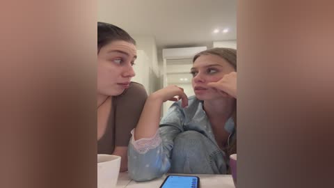 Media: Video of two young women in a casual setting, with one leaning on her hand, and the other looking contemplative, both in light denim outfits, in a modern, minimalistic room with beige walls and recessed lighting.