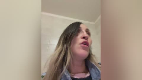 A video of a young woman with long, wavy blonde hair and light skin, wearing a denim jacket, looking upward with a slightly surprised expression. The background shows a beige wall and part of a tiled bathroom.