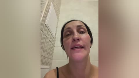 Video of a woman with fair skin and dark hair, smiling in a bathroom with beige and white tiled walls, wearing a black spaghetti strap top.