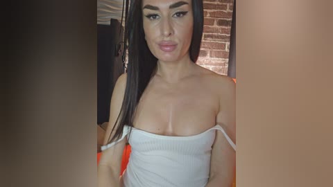 Media: Video of a woman with long, straight black hair, wearing a white, strapless top that reveals her ample cleavage. She has a light skin tone, full lips, and is indoors near a brick wall and hanging garment.