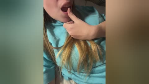 Media: Video of a young woman with fair skin and long, straight blonde hair, wearing a light blue t-shirt, playfully biting her finger. Background is blurred, focusing on her face and upper body.