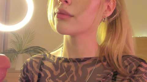 Media: Video of a blonde woman with fair skin, wearing a leopard-print top, and a silver necklace, in a softly lit room with a round ring light in the background.