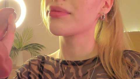 Media: A close-up video of a fair-skinned woman with light blonde hair, wearing a camouflage shirt and dangling earrings, standing indoors with a glowing ring light and a potted palm in the background.