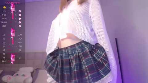 Media: Video of a slender, light-skinned woman in a white, crocheted crop top and a green plaid skirt, posing indoors with a pink teddy bear and a digital time display on the left.