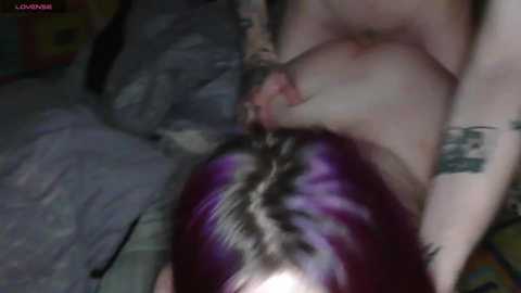 A video depicts a close-up of a naked person with purple hair, lying on a bed, and another person's hand on their chest. The background shows a messy room with a gray blanket.