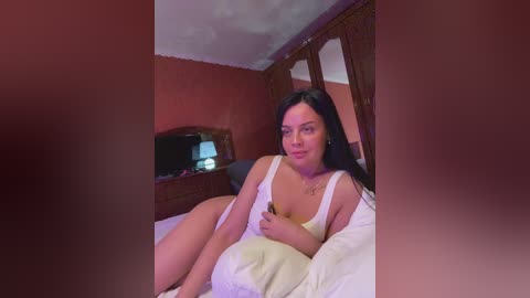 Video of a curvy, light-skinned woman with long black hair, wearing a white tank top, lying on a bed with white sheets in a rustic room with wooden furniture and a TV.