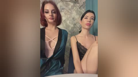 Media: Video of two women with fair skin and dark hair, one in a teal satin robe and the other in black lingerie, sitting on a bed with patterned sheets and teal curtains in the background.