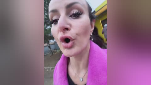 Media: Video of a woman with dark hair, wearing pink coat, looking surprised with open mouth, blurry background with greenery and yellow structure.