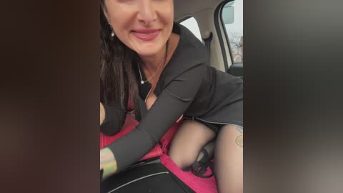 Media: Video of a smiling woman with medium-length dark hair, wearing a black shirt, holding a baby in a red car seat.