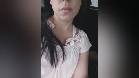 A video of a woman with pale skin and long black hair, wearing a white blouse with a pink bow, seated in a dark car interior.