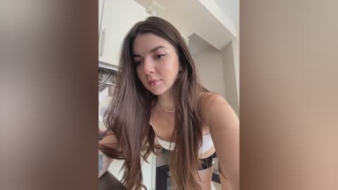 Media: A video of a young woman with long brown hair, wearing a white and black bra, standing in a kitchen with white cabinets and a tiled backsplash.