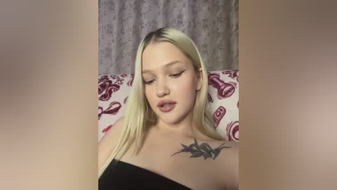 Media: Video of a young woman with long blonde hair, wearing a black top, lying on a bed with a floral patterned sheet. She has a tattoo on her collarbone.