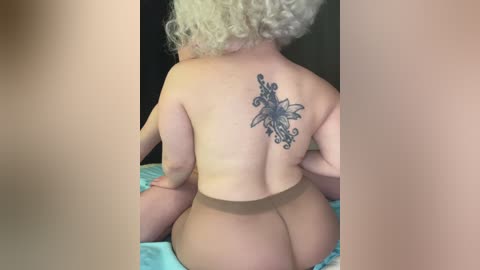 Media: Video of a light-skinned woman with short curly blonde hair, sitting topless, showcasing a large, intricate black butterfly tattoo on her back. She wears sheer, nude-colored tights, highlighting her bare buttocks. The background is dark, focusing attention on her tattoo and figure.