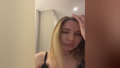Media: Video of a young Caucasian woman with straight, shoulder-length blonde hair, wearing a black lace bra, touching her forehead, in a dimly lit room with beige walls.