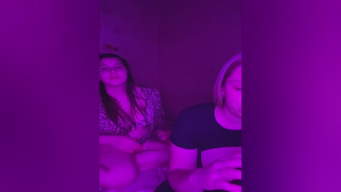 Media: A video with a purple hue shows two women seated, one with long dark hair, the other with blonde hair. They wear casual clothing, seated in a dimly lit room with a low ceiling.