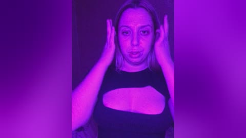 Media: A video of a light-skinned woman with shoulder-length blonde hair, wearing a black crop top with a large cutout, posing against a purple-lit background. She is holding her ears.