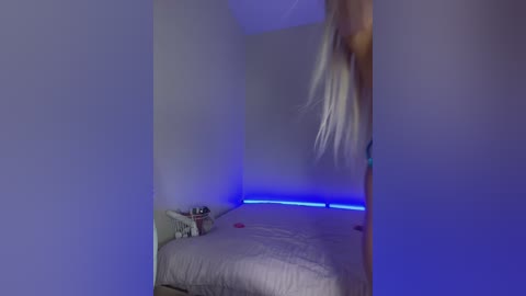 Media: Video of a dimly lit bedroom with a bed and a nightstand; a person with long blonde hair is blurred, possibly dancing. Blue LED lights under the bed create a cool, eerie ambiance.