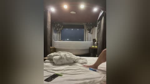 Video of a cozy bedroom with a white round bathtub in the center, dimly lit by ceiling lights, featuring a messy white duvet and a person's leg.