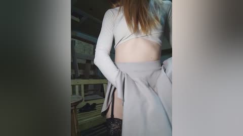 Video of a slender, light-skinned woman with long blonde hair, wearing a white crop top and gray pleated skirt, standing in a dimly lit room with a wooden railing.