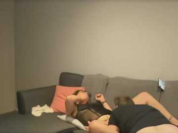 Media: Video of two men lying on a grey sofa in a dimly lit room, one with a shirt on, the other shirtless. A pink pillow and a white blanket are visible.