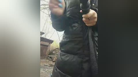 Video of a person in black puffy jacket and gloves, holding a gun, standing in a dilapidated, overgrown yard with wooden structures and trees in the background.