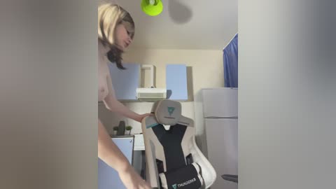 Video of a blonde woman in a beige top adjusting a gaming chair with a grey headrest and black accents in a modern, minimalist room with light blue walls and a green ceiling light.