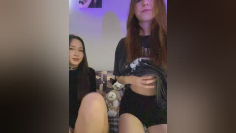 Media: Video of two young women, one with long black hair, wearing a black choker and a black sweater, sitting on a bed with stuffed animals. The other has long brown hair, wearing a black shirt and shorts, holding a shirt.