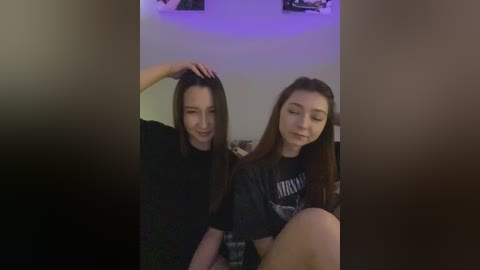 Media: Video of two young women with long hair, one resting her hand on her head, wearing black shirts. Background includes a purple-lit wall and blurred objects.