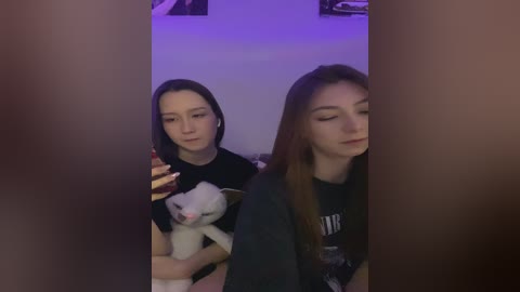 Media: Video of two young women with long brown hair, one holding a white plush toy, in a dimly lit room with purple lighting, both looking relaxed.