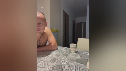 Media: Video of a shirtless, bald Caucasian man leaning on a table with a patterned tablecloth, wearing a necklace, in a modern apartment with white walls and doors.