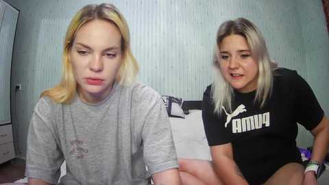 Media: Video of two young women with blonde hair, one wearing a grey T-shirt, the other a black Puma shirt. They sit on a bed in a simple room with light blue walls and a white wardrobe.