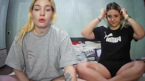 Media: Video of two young women, one with blonde hair, one with brown hair, both wearing Puma t-shirts, sitting cross-legged on a bed in a dimly lit, minimalistic room.