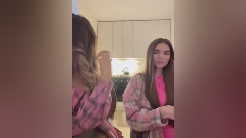 Media: A video of a young woman with long, straight brown hair, wearing a pink plaid shirt and pink top, sitting on a chair in a kitchen with white cabinets and a sink.