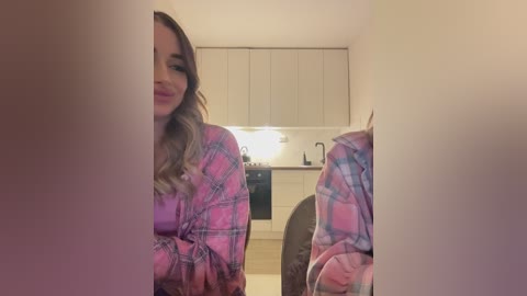 Media: Video of a young woman with light skin, wavy brown hair, wearing a pink plaid pajama shirt, sitting in a modern, well-lit kitchen with white cabinets and a stainless steel oven.