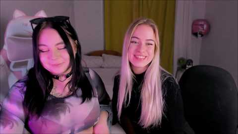 Video of two smiling young women in cat costumes with black and white fur, one with long black hair, the other with long blonde hair, in a dimly lit bedroom with a bed and yellow curtains.