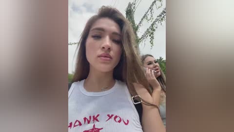 Media: Video of two young women, one with long brown hair wearing a white tank top with \"THANK YOU\" in red, standing outdoors near trees, blurred background.