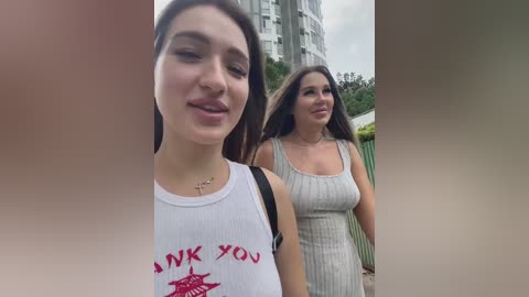 Media: Video of two young women, one in a white tank top with a red \"THANK YOU\" graphic, the other in a ribbed grey dress, walking outdoors in front of a modern high-rise building.