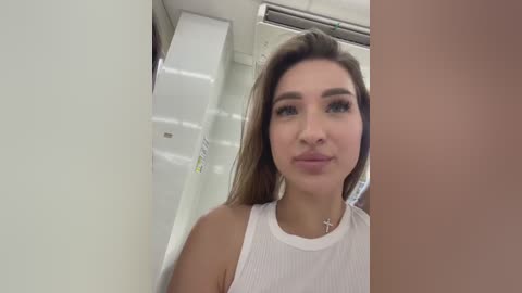 Media: Video of a young woman with straight brown hair and light skin, wearing a white sleeveless top, standing in a medical setting with white walls and a ceiling air vent.