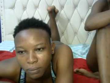 Video of a Black woman with short hair lying on a red bedspread in a bedroom, wearing a black and white striped bra, against a tufted white headboard.
