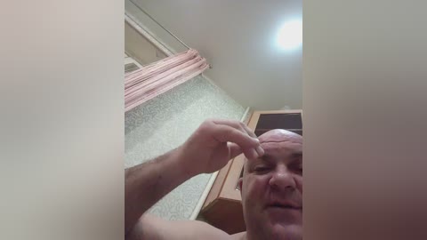 A close-up video of an elderly man with a bald head, light skin, and a wrinkled face, combing his hair in a bathroom with light green wallpaper and a pink curtain.