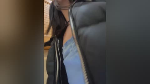 Media: Video of a person wearing a black puffer jacket, light blue jeans, and a black choker, standing indoors near a window with blinds.