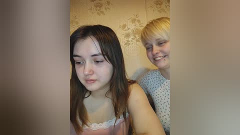 Media: Video of two young girls, one with long dark hair in a pink dress, the other with short blonde hair in a blue dress, standing side by side against a floral wallpaper background.