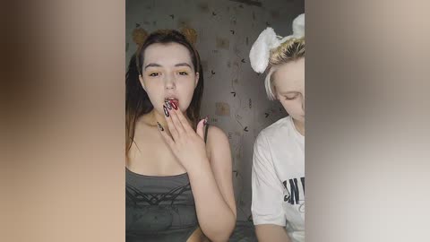 Media: Video of two young women in a dimly lit room. One with long brown hair, wearing a gray top, licking her fingers, while the other, with short blonde hair and wearing a white t-shirt, stands to the right.