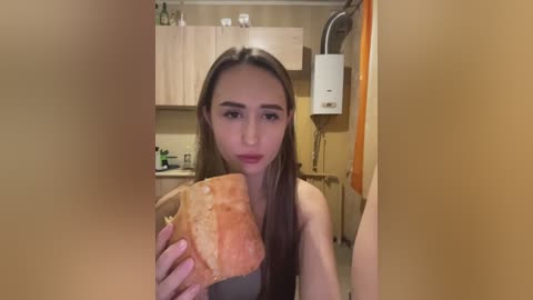 Video of a young woman with straight brown hair, fair skin, and a neutral expression, holding a partially eaten sandwich. She's in a small, cluttered kitchen with light wood cabinets, a white water heater, and a hanging orange towel.